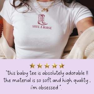 May include: White t-shirt with a maroon graphic of cowboy boots and the text "SAVE A HORSE". The t-shirt is being worn by a person. The review text reads "this baby tee is absolutely adorable !! the material is so soft and high quality. i'm obsessed."