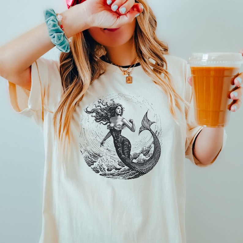 May include: A white t-shirt with a black and white illustration of a mermaid with long flowing hair and a tail. The mermaid is surrounded by waves and the illustration is in a circle. The t-shirt is being worn by a person with a pink headband and a gold necklace with a small letter 'A' pendant.
