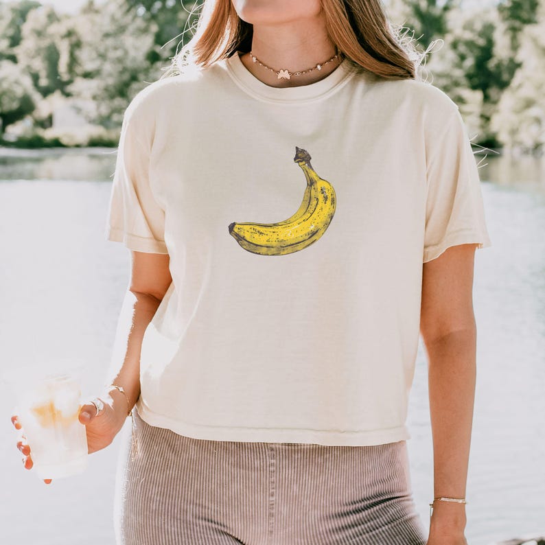 May include: Cream-colored t-shirt featuring a yellow banana graphic. The banana is depicted in a detailed, illustrative style. The shirt has short sleeves and a relaxed fit, suitable for casual wear.