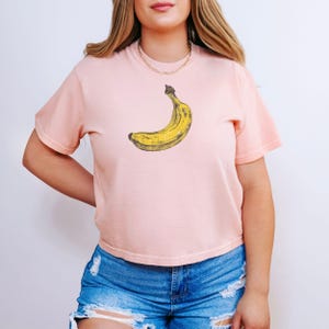 May include: A light pink t-shirt featuring a yellow banana graphic. The banana is slightly curved and has a brown stem. The shirt has short sleeves and is paired with blue denim shorts.