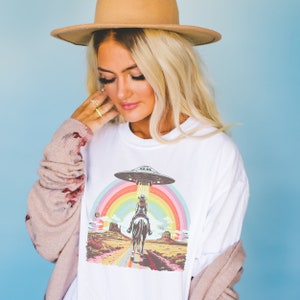 Op de afbeelding: Wit T-shirt met een afbeelding van een vrouw die op een paard rijdt met een regenboog en een UFO erboven. De afbeelding is geplaatst tegen een woestijnlandschap.