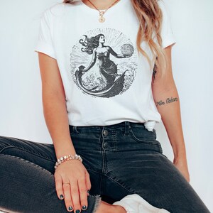 May include: White t-shirt with a black and white illustration of a mermaid holding a globe. The mermaid is surrounded by waves and the text "The Ocean" is visible on the right side of the image.