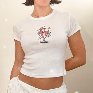 May include: White, short-sleeved t-shirt featuring a cartoon bull character in a martini glass. The bull is red and the martini glass is black and white. The shirt is a fitted style.