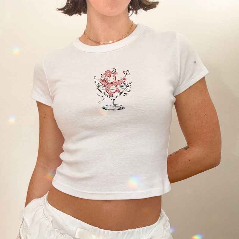 May include: White, short-sleeved t-shirt featuring a cartoon bull character in a martini glass. The bull is red and the martini glass is black and white. The shirt is a fitted style.