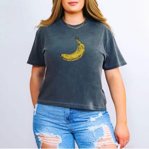 May include: Dark gray t-shirt featuring a yellow banana graphic. The banana is positioned on the front of the shirt. The shirt has short sleeves and a crew neck. The model is wearing blue jeans.