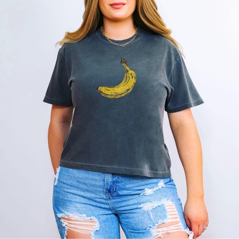 May include: Dark gray t-shirt featuring a yellow banana graphic. The banana is positioned on the front of the shirt. The shirt has short sleeves and a crew neck. The model is wearing blue jeans.
