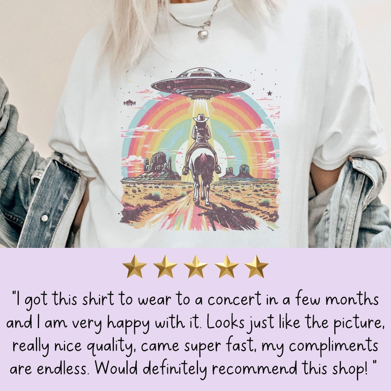 Op de afbeelding: Een wit T-shirt met een grafische print van een cowboy die op een paard rijdt in een woestijnlandschap met een regenboog en een UFO erboven. De tekst op het T-shirt luidt "I got this shirt to wear to a concert in a few months and I am very happy with it. Looks just like the picture, really nice quality, came super fast, my compliments are endless. Would definitely recommend this shop!"