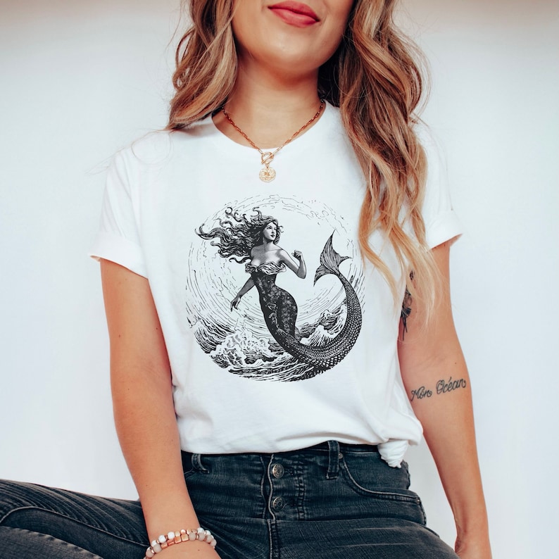 May include: White t-shirt with a black and white illustration of a mermaid with long flowing hair, a tail, and a crown. The mermaid is surrounded by waves and the text "Free Ocean".