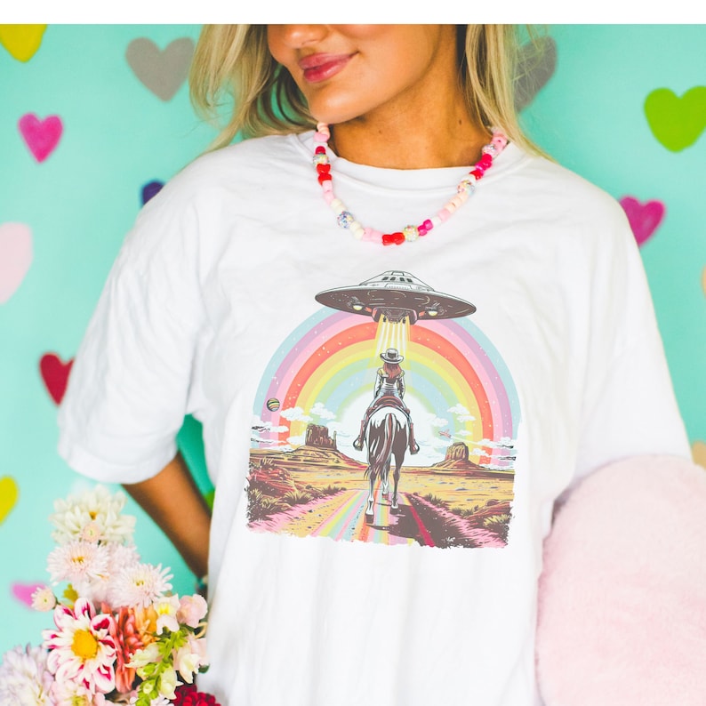 Op de afbeelding: Wit T-shirt met een afbeelding van een cowgirl die een paard rijdt onder een regenboog met een UFO erboven.