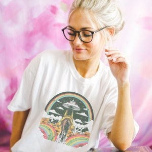 May include: White t-shirt with a graphic of a woman riding a horse through a rainbow with a UFO in the sky. The graphic is in a retro style with a black background and colorful accents.