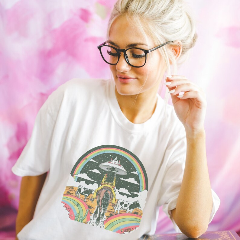 May include: White t-shirt with a graphic of a woman riding a horse through a rainbow with a UFO in the sky. The graphic is in a retro style with a black background and colorful accents.