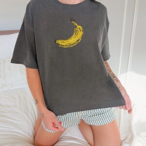 May include: Dark gray t-shirt featuring a yellow banana graphic. The shirt has a relaxed fit and short sleeves. The person is wearing striped shorts. The shirt is made of a soft material.