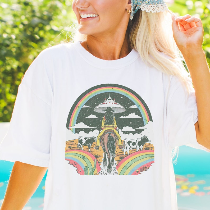 May include: White t-shirt with a graphic of a woman riding a horse through a rainbow with a UFO in the background. The woman is wearing a cowboy hat and a yellow shirt. There are two cows in the background.