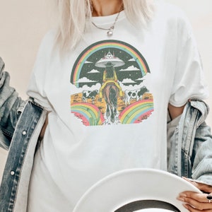 May include: A white t-shirt with a graphic print of a cowgirl riding a horse under a rainbow with a UFO in the sky. The graphic is in a vintage style with a black background and colorful accents.