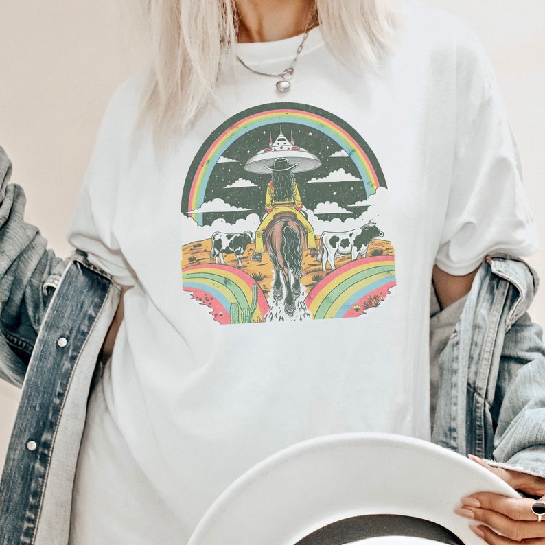 May include: A white t-shirt with a graphic print of a cowgirl riding a horse under a rainbow with a UFO in the sky. The graphic is in a vintage style with a black background and colorful accents.