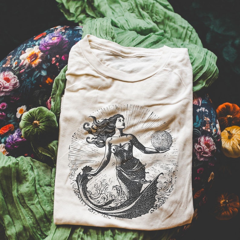 May include: A white t-shirt with a black and white illustration of a mermaid holding a sphere. The mermaid has long flowing hair and is wearing a shell bra. The illustration is surrounded by a circle of waves.