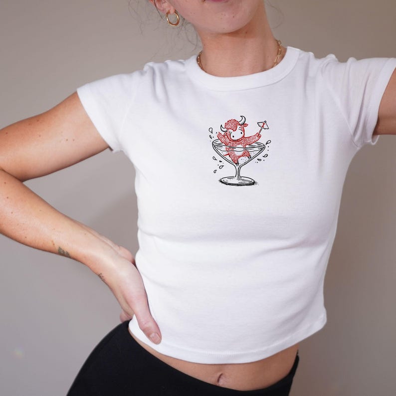 May include: White crop top featuring a whimsical illustration of a red bull character relaxing in a martini glass. The bull is holding a tiny cocktail umbrella. The graphic is in black and red. The shirt has short sleeves and a crew neck.