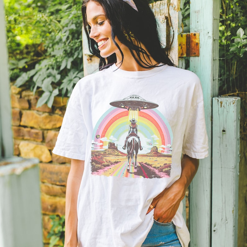 Op de afbeelding: Een wit T-shirt met een afbeelding van een cowgirl die op een paard door een regenboog rijdt met een UFO erboven. De afbeelding is in retro-stijl.