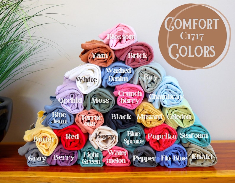 Op de afbeelding: Een stapel gerolde stofstalen in verschillende kleuren, waaronder blauw, groen, rood, geel en roze. De stofstalen zijn gelabeld met kleurnamen, zoals "Blue Jean", "Red", "Butter" en "Berry". De tekst "COMFORT C1717 COLORS" is zichtbaar bovenaan de afbeelding.