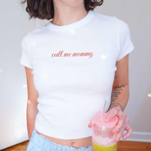 May include: White, short-sleeved t-shirt with the phrase "call me mommy" in red script. The shirt is a fitted style. The person is holding a pink and clear cup with a straw and a green beverage.
