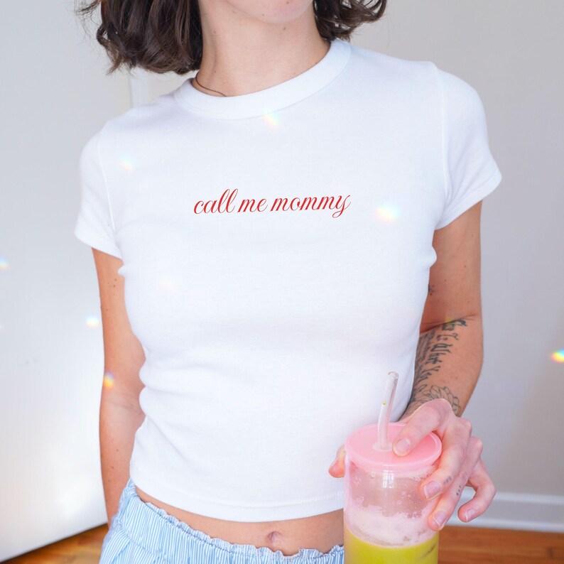 May include: White, short-sleeved t-shirt with the phrase "call me mommy" in red script. The shirt is a fitted style. The person is holding a pink and clear cup with a straw and a green beverage.
