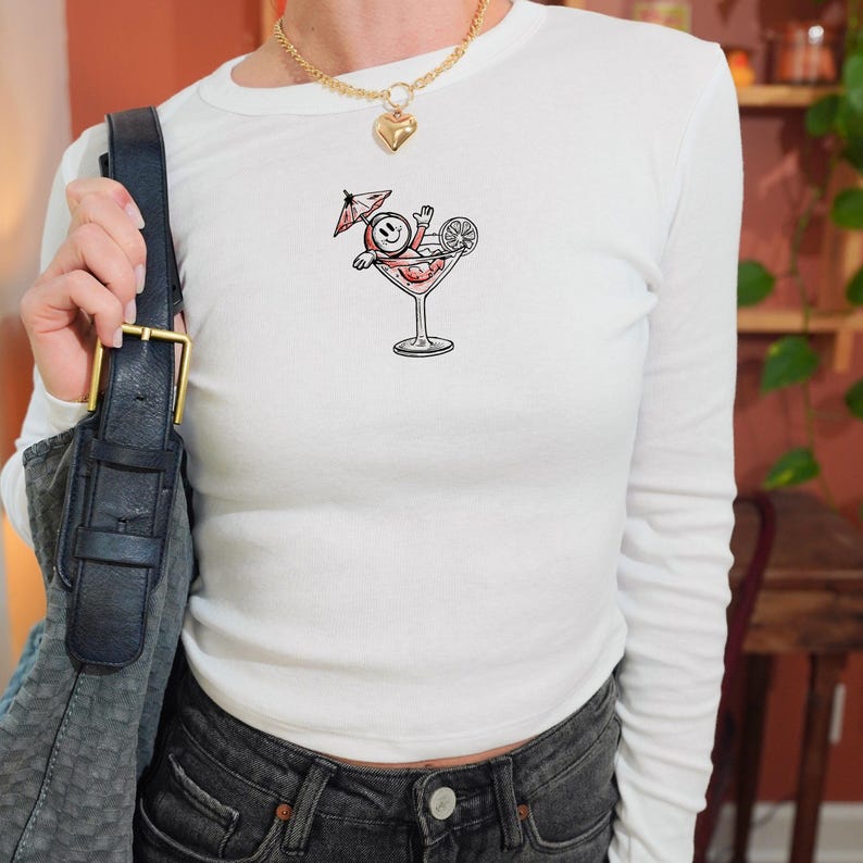 May include: White long-sleeved top featuring a cocktail graphic. The graphic shows a smiling face relaxing in a martini glass with a cocktail umbrella and a lemon slice. The top is worn with dark denim jeans and a black handbag.