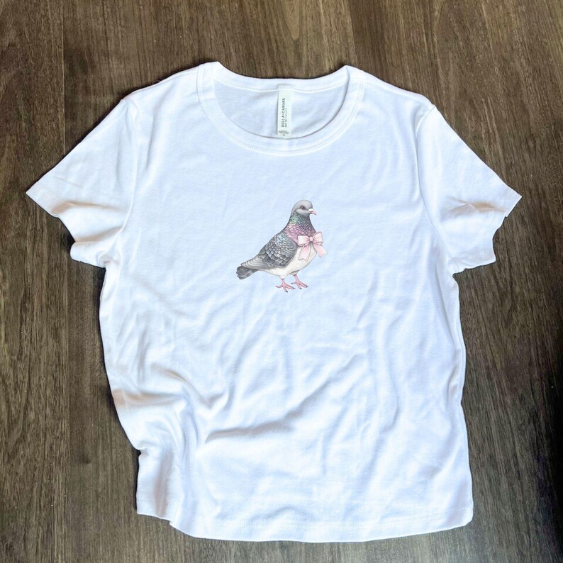 Puede incluir: Camiseta blanca con un gr&aacute;fico de una paloma con un lazo rosa.