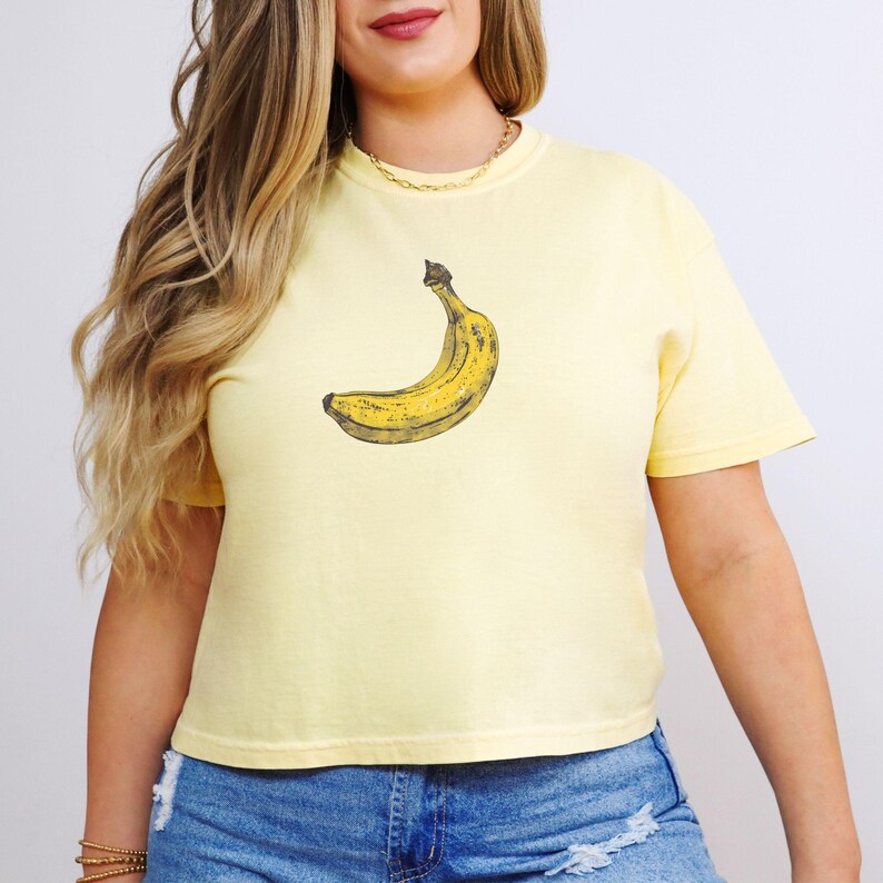 May include: A light yellow t-shirt featuring a detailed illustration of a banana. The banana is yellow with brown shading. The shirt has short sleeves and a crew neck. The model is wearing blue denim shorts and a gold chain.