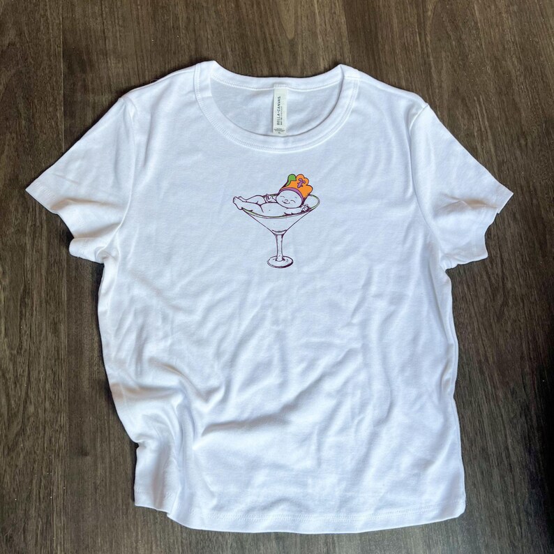 May include: White t-shirt with a graphic of a baby wearing a crown and lying in a martini glass.