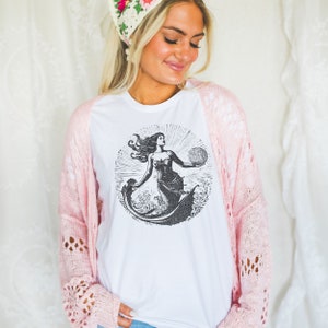 May include: White t-shirt with a black and white illustration of a mermaid holding a seashell. The mermaid is surrounded by waves and the sun is shining behind her.