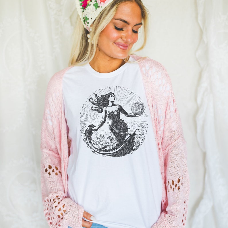 May include: White t-shirt with a black and white illustration of a mermaid holding a seashell. The mermaid is surrounded by waves and the sun is shining behind her.