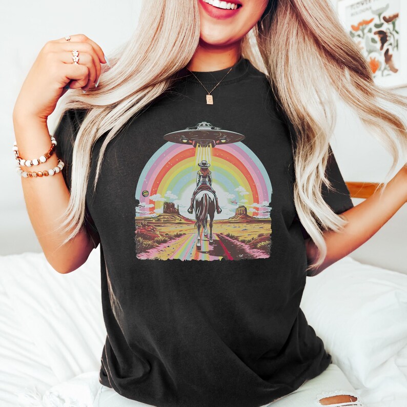 Op de afbeelding: Een zwart T-shirt met een afbeelding van een cowgirl die op een paard door een regenboog rijdt met een UFO erboven. De afbeelding is in retro-stijl.