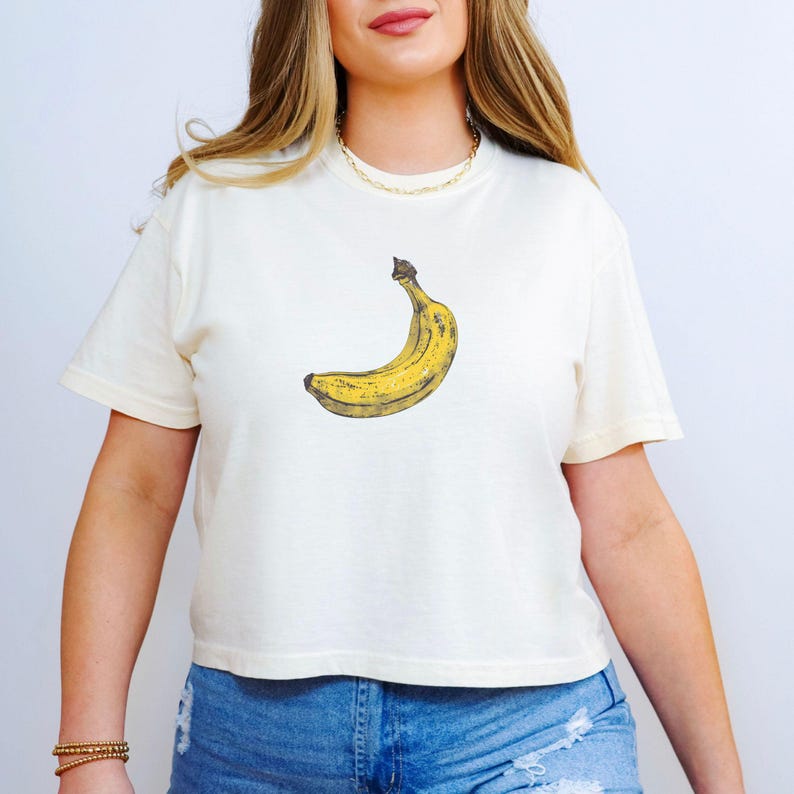 May include: Cream-colored t-shirt featuring a yellow banana graphic. The banana is depicted in a realistic style with brown and yellow hues. The shirt has short sleeves and a crew neck.