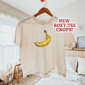 May include: Cream-colored boxy tee shirt with a yellow banana graphic. The shirt is hanging on a wooden hanger. Text on the shirt reads "NEW BOXY TEE CROPS!"