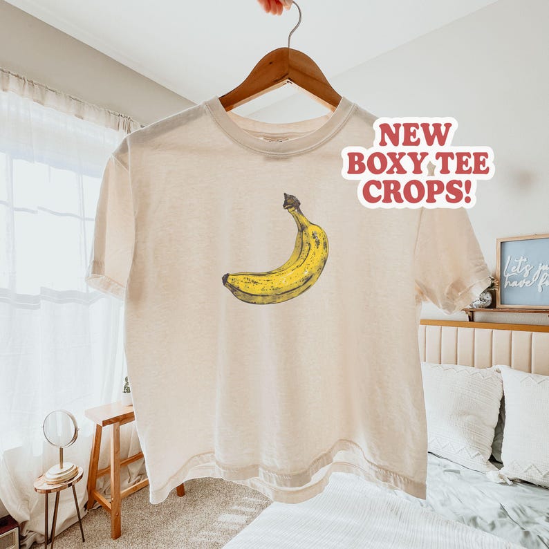 May include: Cream-colored boxy tee shirt with a yellow banana graphic. The shirt is hanging on a wooden hanger. Text on the shirt reads "NEW BOXY TEE CROPS!"