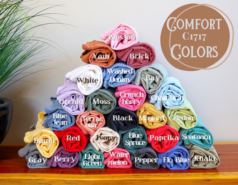 May include: A stack of rolled t-shirts in various colors, including white, red, blue, green, and yellow. The t-shirts are labeled with names like "Blossom", "Yam", "Washed Denim", "Brick", "Bay", "Orchid", "Moss", "Crunch Berry", "Black", "Mustard", "Celadon", "Blue Jean", "Butter", "Red", "Ivory", "Blue Spruce", "Paprika", "Seafoam", "Gray", "Berry", "Light Green", "Watermelon", "Pepper", "Flo Blue", and "Khaki". The text "COMFORT C1717 COLORS" is visible in the top right corner of the image.