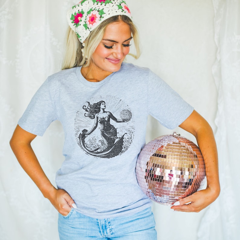 May include: A gray t-shirt with a black and white illustration of a mermaid holding a globe. The mermaid is surrounded by waves. The shirt is being held by a person wearing a white crocheted hat with pink flowers.