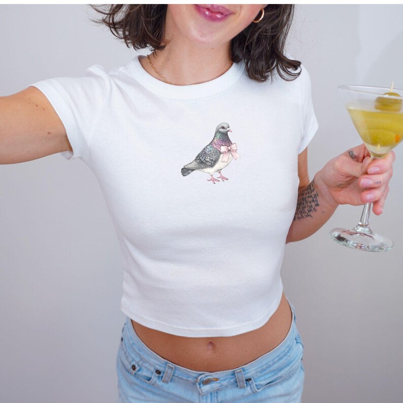 Puede incluir: Camiseta blanca recortada con un gr&aacute;fico de una paloma con un lazo rosa.