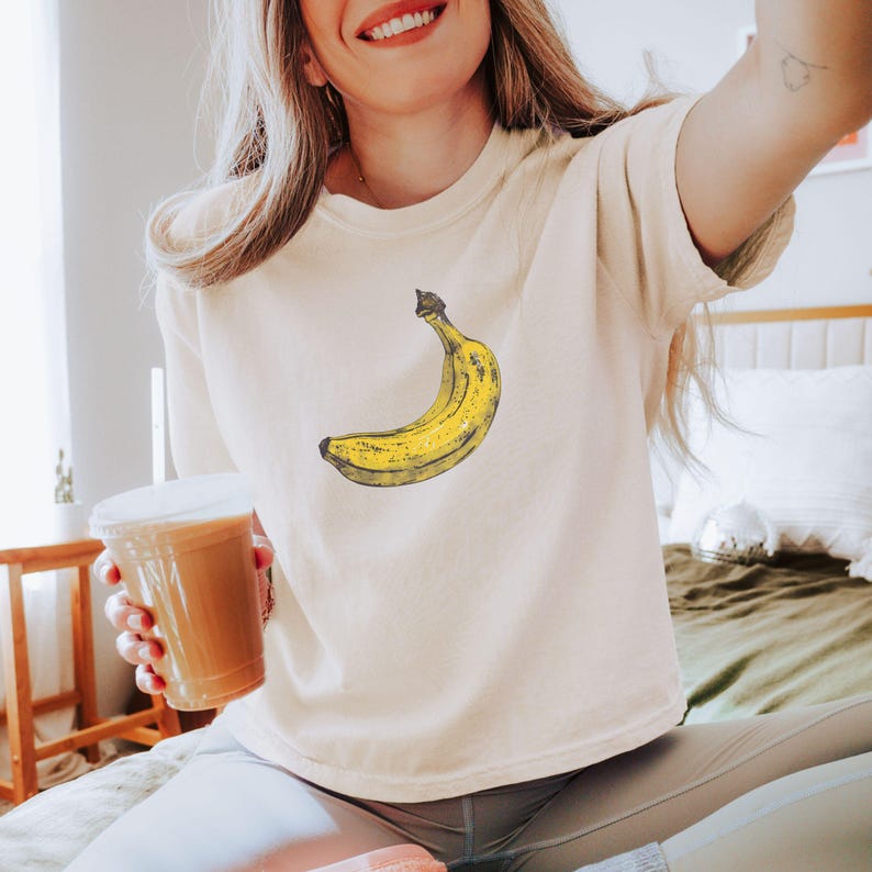 May include: Cream-colored t-shirt featuring a yellow banana graphic. The banana is drawn in a vintage style with black outlines and shading. The shirt has a classic crew neck and short sleeves, made of a soft, comfortable material.