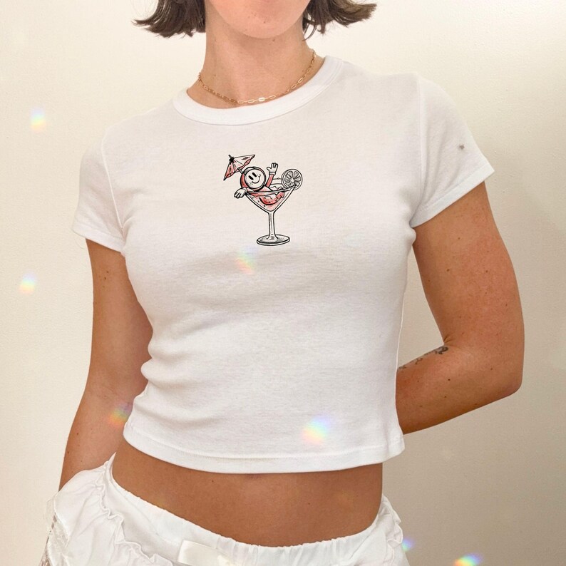 May include: White, short-sleeved crop top with a round neckline. The shirt features a graphic of a smiling face with an umbrella in a martini glass. The graphic is in red, black, and white. The shirt is made of a soft, ribbed material.