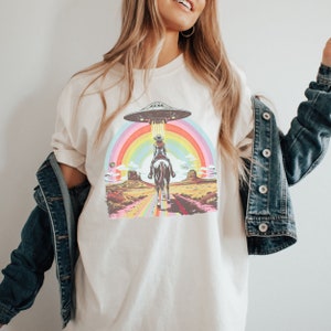 Op de afbeelding: Een wit T-shirt met een afbeelding van een vrouw die op een paard door een regenboog rijdt met een UFO erboven. De afbeelding is in retro-stijl.