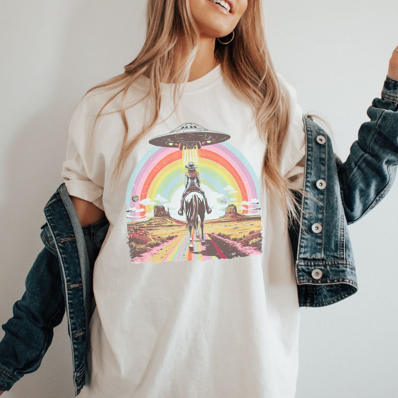 Op de afbeelding: Een wit T-shirt met een afbeelding van een vrouw die op een paard door een regenboog rijdt met een UFO erboven. De afbeelding is in retro-stijl.