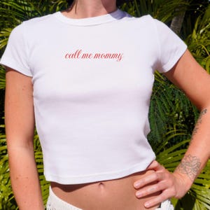 May include: White, short-sleeved crop top with the phrase "call me mommy" in red script across the chest. The shirt has a crew neck and is made of a soft material. Green foliage forms the background.