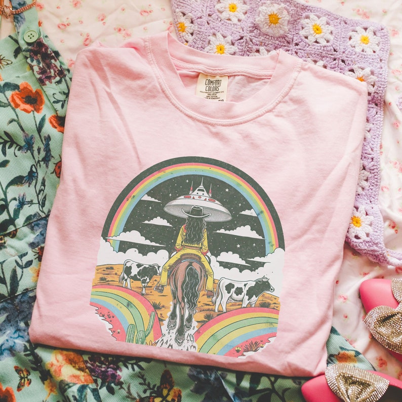 May include: Pink t-shirt with a graphic of a woman riding a horse through a rainbow with a UFO in the background. The text "COMFORT COLORS" is on the tag.