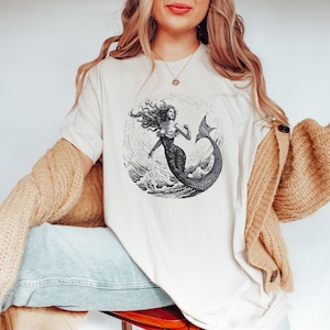 May include: A white t-shirt with a black and white illustration of a mermaid in a circle. The mermaid has long flowing hair and is holding her tail up.