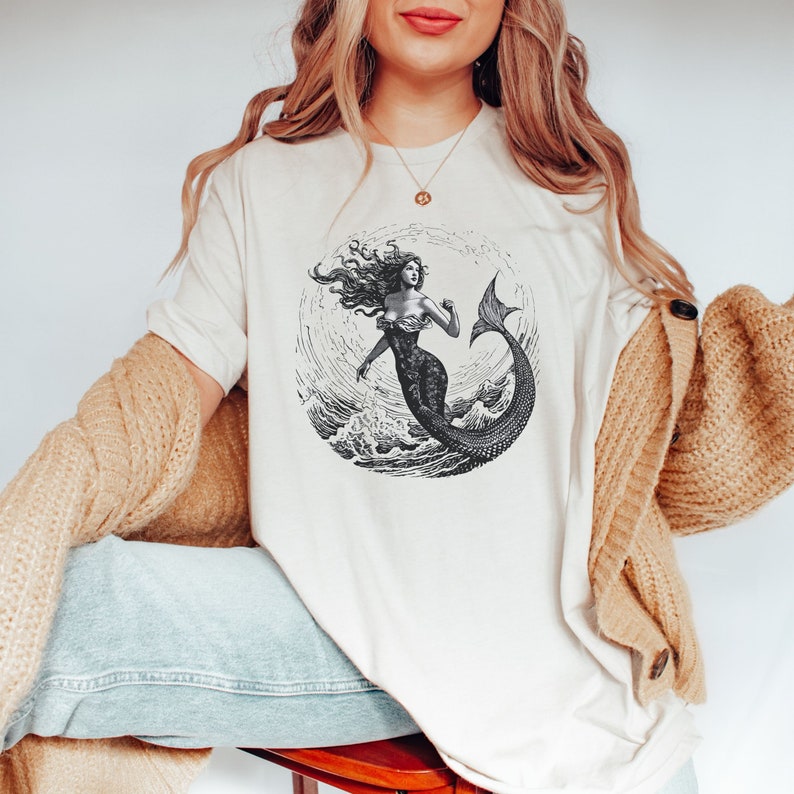 May include: A white t-shirt with a black and white illustration of a mermaid in a circle. The mermaid has long flowing hair and is holding her tail up.