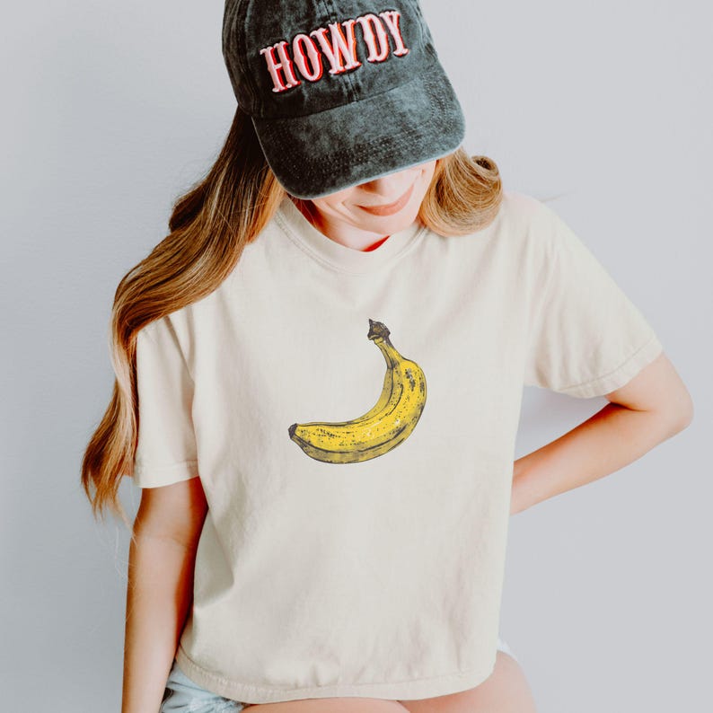 May include: Cream-colored t-shirt featuring a yellow banana graphic. The person is wearing a dark green cap with the word "HOWDY" in red letters. The shirt has short sleeves.
