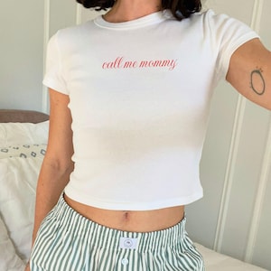 May include: A white crop top with short sleeves, featuring the red text "call me mommy". The top is worn with green and white striped shorts. The image presents a relaxed, everyday style.