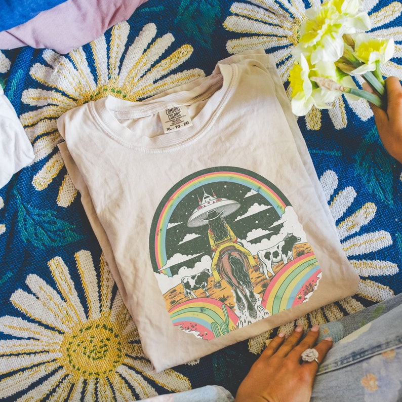 May include: A beige t-shirt with a graphic print of a woman riding a horse through a rainbow with a UFO in the background. The text "Cosmic Colors" and "XL - TO - BIG" is printed on the tag.