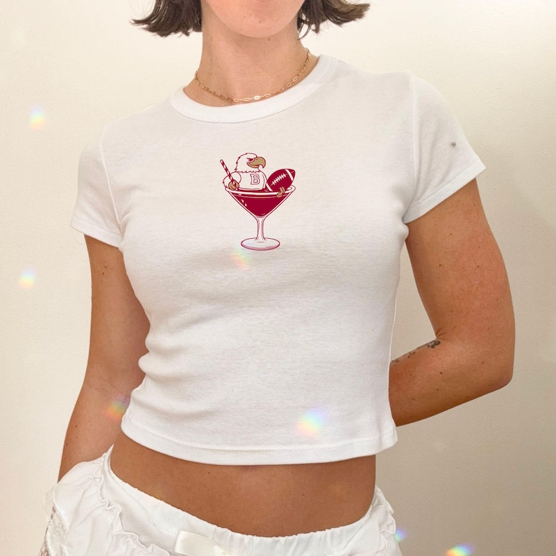 May include: White, short-sleeved t-shirt with a maroon graphic of an eagle, football, and the letter "B" in a martini glass. The shirt has a fitted shape and a crew neck.